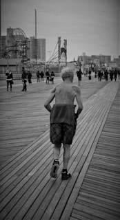 An elderly man jogs shirtless down a wooden boardwalk surrounded by people walking in both directions. The background features an urban skyline with high-rise buildings and amusement park rides. The scene is captured in black and white, creating a nostalgic atmosphere.
