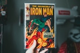 A person is holding a vintage comic book of 'The Invincible Iron Man'. The cover art features Iron Man in a dramatic scene with flames in the background and another character in a green outfit. The comic is enclosed in a protective plastic casing.