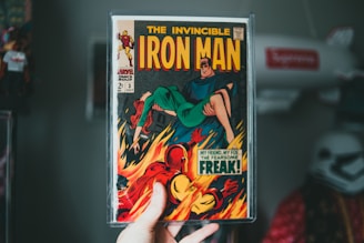 A person is holding a vintage comic book of 'The Invincible Iron Man'. The cover art features Iron Man in a dramatic scene with flames in the background and another character in a green outfit. The comic is enclosed in a protective plastic casing.