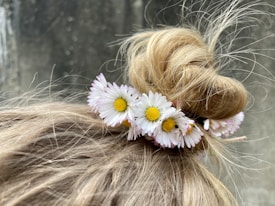 A close-up view of a hair bun adorned with small white and yellow daisies. The hair is a light blonde color, and loose strands are visible, giving a casual and natural appearance. The background is blurred, emphasizing the floral arrangement in the hair.
