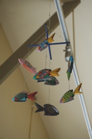 A colorful mobile featuring various fish-shaped ornaments hanging from strings. Each fish is uniquely decorated with vibrant patterns and colors, and they are arranged in a circular fashion around a central axis. The background shows the ceiling and parts of structural beams, giving an impression of an indoor setting.