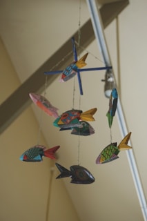 A colorful mobile featuring various fish-shaped ornaments hanging from strings. Each fish is uniquely decorated with vibrant patterns and colors, and they are arranged in a circular fashion around a central axis. The background shows the ceiling and parts of structural beams, giving an impression of an indoor setting.