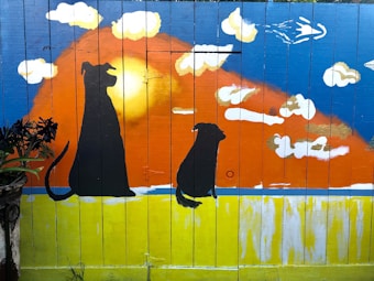 A vibrant mural painted on a wooden fence features two silhouetted dogs facing a dramatic sunset. The sky transitions from deep blue to orange, surrounded by fluffy white clouds. A bird flies across the sky, and a plant is visible on the left side of the image.