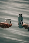 Two hands are extending towards each other, one holding a white enamel mug with a deer silhouette, and the other holding a silver and teal thermos with a logo. The background features a blurred body of water, suggesting an outdoor setting.