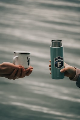 Two hands are extending towards each other, one holding a white enamel mug with a deer silhouette, and the other holding a silver and teal thermos with a logo. The background features a blurred body of water, suggesting an outdoor setting.