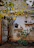 A rustic wall adorned with decorative nautical items like ship wheels, an anchor, and vintage fishing net. Two shelves display various items including old kettles and gourds. The area is surrounded by green and yellow foliage, giving a natural and quaint charm.