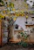A rustic wall adorned with decorative nautical items like ship wheels, an anchor, and vintage fishing net. Two shelves display various items including old kettles and gourds. The area is surrounded by green and yellow foliage, giving a natural and quaint charm.