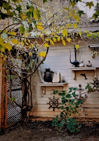 A rustic wall adorned with decorative nautical items like ship wheels, an anchor, and vintage fishing net. Two shelves display various items including old kettles and gourds. The area is surrounded by green and yellow foliage, giving a natural and quaint charm.