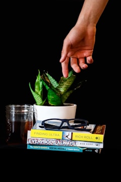 A hand reaching out over a small stack of books placed next to a potted plant and a glass jar with iced tea. The books include titles like 'Finding Ultra' and 'Everybody Always' with a pair of dark-framed glasses resting on top.