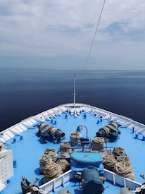 The deck of a large ship is visible, painted bright blue and surrounded by calm, open water. Numerous coils of rope are arranged neatly along the deck, along with various marine equipment. The horizon blends with a slightly overcast sky, creating a tranquil and expansive seascape.