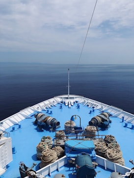 The deck of a large ship is visible, painted bright blue and surrounded by calm, open water. Numerous coils of rope are arranged neatly along the deck, along with various marine equipment. The horizon blends with a slightly overcast sky, creating a tranquil and expansive seascape.