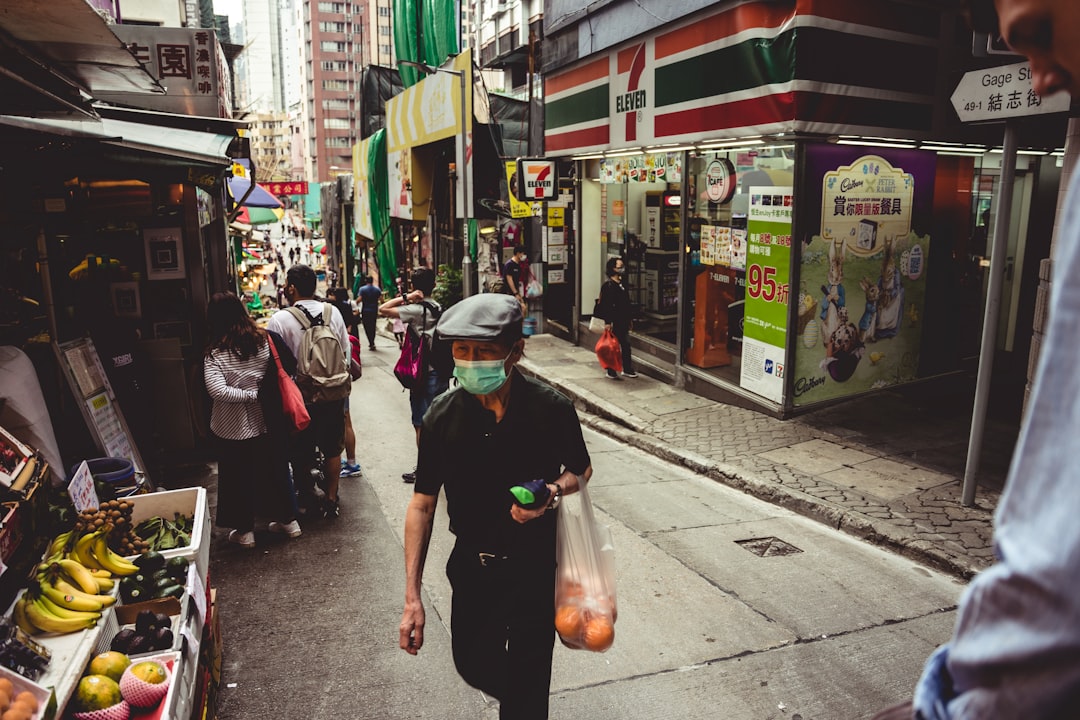 man in black tank top and white cap holding orange plastic bag, 3:43PM - cruising in HK market street 🇭🇰🍊