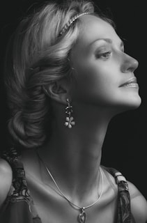 A black-and-white portrait of a woman in profile, with elegantly styled hair and adorned with a headband. She wears dangling earrings and a necklace with a heart-shaped pendant. Her expression is serene and poised.