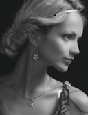 A woman with styled hair is adorned with a braided headband and wears dangling earrings with floral motifs. She sports a heart-shaped pendant necklace and has an air of elegance. Her attire includes patterned fabric visible on her shoulder.