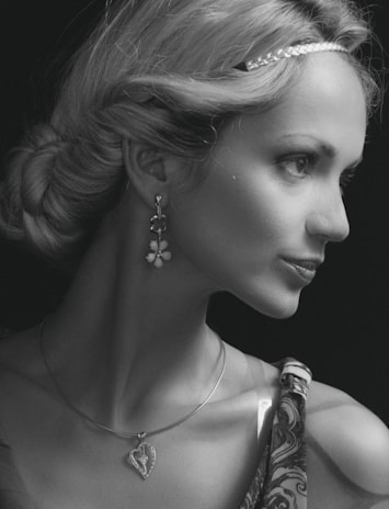 A woman with styled hair is adorned with a braided headband and wears dangling earrings with floral motifs. She sports a heart-shaped pendant necklace and has an air of elegance. Her attire includes patterned fabric visible on her shoulder.