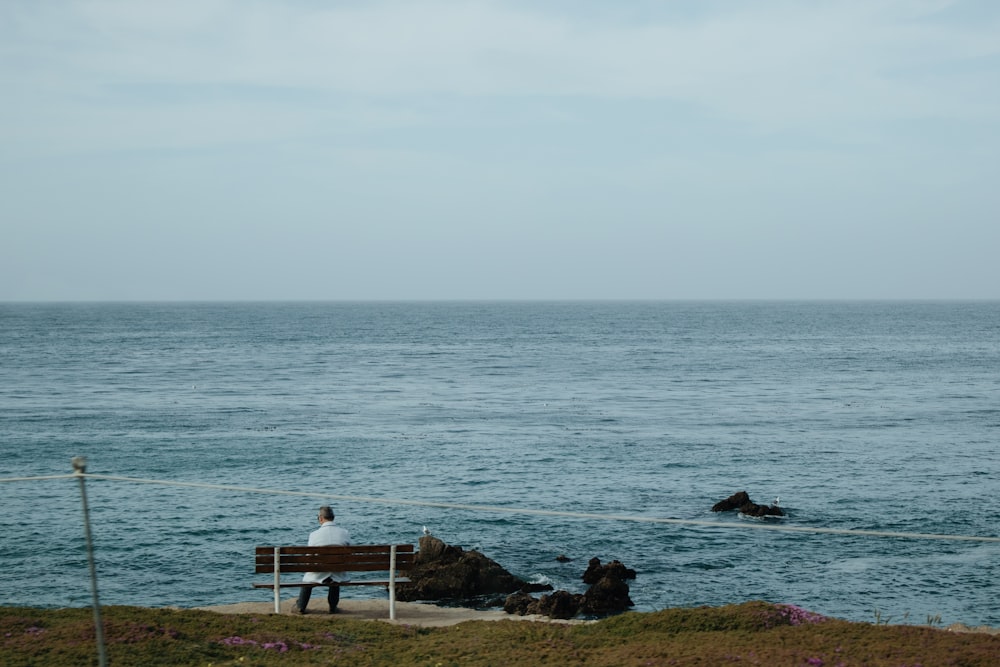 person sitting on white wooden bench near sea during daytime