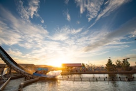 A water ride splashes down with a large plume of water under a bright sunset. The sky is vast and partly cloudy, with a warm glow from the sun casting light across the amusement park area. People are likely on the ride, and there are trees and structures in the background.
