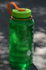 A green plastic water bottle with measurement markings on its side sits on a flat surface. The bottle has a bright green lid with an orange handle attached. The image is lit with natural daylight, casting soft shadows on the surface.