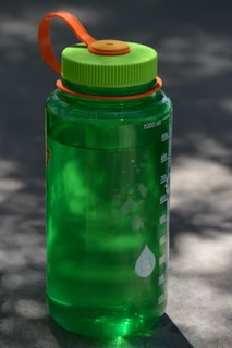A green plastic water bottle with measurement markings on its side sits on a flat surface. The bottle has a bright green lid with an orange handle attached. The image is lit with natural daylight, casting soft shadows on the surface.