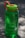 A green plastic water bottle with measurement markings on its side sits on a flat surface. The bottle has a bright green lid with an orange handle attached. The image is lit with natural daylight, casting soft shadows on the surface.