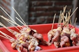 Several skewers of raw meat and organs, possibly prepared for a barbecue, are arranged on a red tray. The skewers contain different types of meat, including chunks of red meat and yellow fat, indicating a variety of cuts.