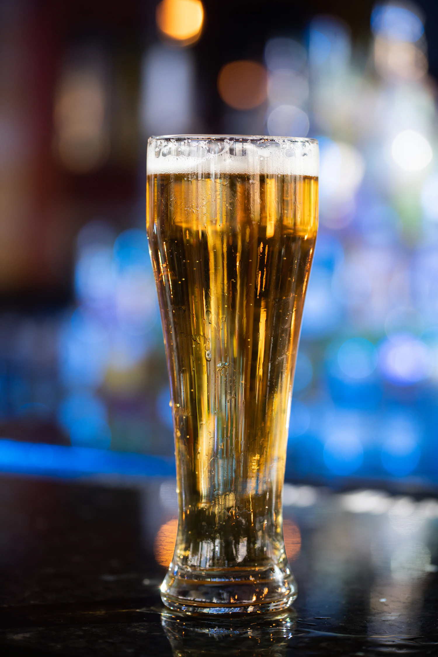 A tall pilsner glass of sparkling pale lager with a white head, blue bar lights behind