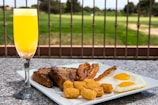 A breakfast plate featuring sunny-side-up eggs, crispy bacon strips, golden-brown tater tots, and thick slices of toast placed on a granite surface. A glass of orange juice is next to the plate. In the background, a landscaped garden is visible, separated by a wrought iron fence.