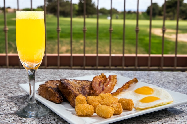A breakfast plate featuring sunny-side-up eggs, crispy bacon strips, golden-brown tater tots, and thick slices of toast placed on a granite surface. A glass of orange juice is next to the plate. In the background, a landscaped garden is visible, separated by a wrought iron fence.