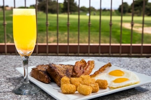 A breakfast plate featuring sunny-side-up eggs, crispy bacon strips, golden-brown tater tots, and thick slices of toast placed on a granite surface. A glass of orange juice is next to the plate. In the background, a landscaped garden is visible, separated by a wrought iron fence.