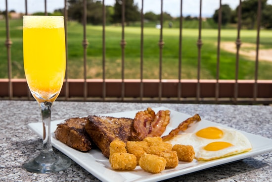 A breakfast plate featuring sunny-side-up eggs, crispy bacon strips, golden-brown tater tots, and thick slices of toast placed on a granite surface. A glass of orange juice is next to the plate. In the background, a landscaped garden is visible, separated by a wrought iron fence.