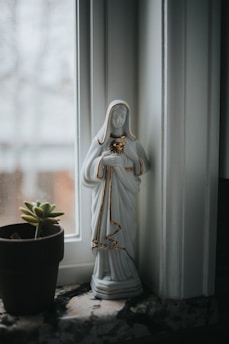 A serene statue of a religious figure in white robes with gold accents stands on a windowsill next to a small succulent plant in a brown pot. Natural light from the window illuminates the scene, creating a peaceful atmosphere.