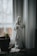 A serene statue of a religious figure in white robes with gold accents stands on a windowsill next to a small succulent plant in a brown pot. Natural light from the window illuminates the scene, creating a peaceful atmosphere.