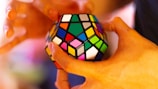 Hands are holding a colorful dodecahedron puzzle with various colored squares on each face. The background is softly blurred, emphasizing the puzzle. The person appears to be in the process of manipulating or solving the puzzle.