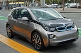 A compact, modern electric car with a sleek design parked on the side of a city street. The vehicle features a two-tone color scheme of gray and black with blue accents on the front grille, and stylish geometric rims. Surrounding the car is an urban setting with markings on the road and blurred buildings in the background.