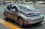 A compact, modern electric car with a sleek design parked on the side of a city street. The vehicle features a two-tone color scheme of gray and black with blue accents on the front grille, and stylish geometric rims. Surrounding the car is an urban setting with markings on the road and blurred buildings in the background.