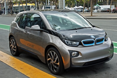 A compact, modern electric car with a sleek design parked on the side of a city street. The vehicle features a two-tone color scheme of gray and black with blue accents on the front grille, and stylish geometric rims. Surrounding the car is an urban setting with markings on the road and blurred buildings in the background.