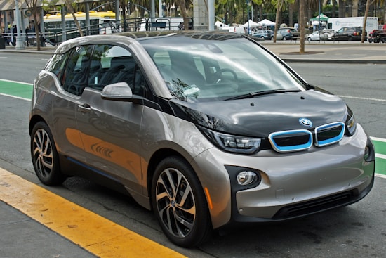 A compact, modern electric car with a sleek design parked on the side of a city street. The vehicle features a two-tone color scheme of gray and black with blue accents on the front grille, and stylish geometric rims. Surrounding the car is an urban setting with markings on the road and blurred buildings in the background.