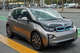A compact, modern electric car with a sleek design parked on the side of a city street. The vehicle features a two-tone color scheme of gray and black with blue accents on the front grille, and stylish geometric rims. Surrounding the car is an urban setting with markings on the road and blurred buildings in the background.