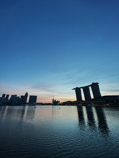 The image captures a tranquil cityscape during dusk or dawn. The skyline features a distinctive hotel with three towers and a ship-like structure on top, situated next to a calm body of water. Reflections of the buildings are visible on the water's surface, and the sky displays a gradient of blue transitioning to orange and pink near the horizon.