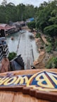A water ride is seen from a high vantage point, surrounded by lush greenery and rustic buildings. The track leads towards a pool of water, and there are people walking along a pathway adjacent to the ride. The atmosphere appears to be that of an amusement park.