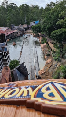 A water ride is seen from a high vantage point, surrounded by lush greenery and rustic buildings. The track leads towards a pool of water, and there are people walking along a pathway adjacent to the ride. The atmosphere appears to be that of an amusement park.