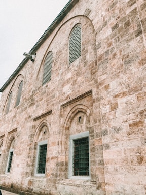 A historic stone building characterized by arched windows and intricate stonework. The structure features a series of evenly spaced windows with protective grilles, and the stone surface has a warm, textured appearance suggesting age and antiquity.