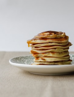 A stack of fluffy pancakes drizzled with syrup, set against a backdrop of burnt orange and cream beige tones.
