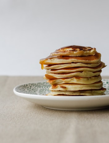 A stack of fluffy pancakes drizzled with syrup, set against a backdrop of burnt orange and cream beige tones.