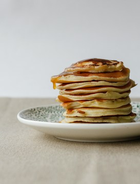 A stack of fluffy pancakes is placed on a decorative plate. The pancakes are generously topped with a golden drizzle of syrup, creating an appetizing appearance. The scene is set against a neutral background with a simple tablecloth, emphasizing the focus on the pancakes.