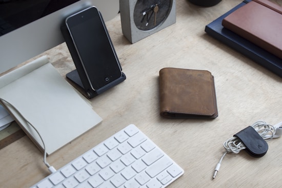A neatly arranged workspace featuring a wooden desk with a notepad, smartphone on a stand, leather wallet, coiled earphones secured with a clip, and a compact keyboard.
