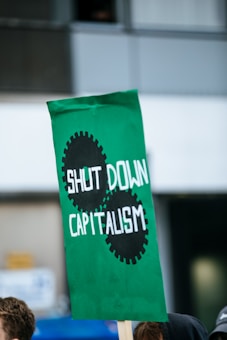 A green protest sign with black gears depicting the words 'SHUT DOWN CAPITALISM.' The sign has a wrinkled appearance and is held by an unseen person in a public setting with blurred windows in the background.