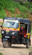 A black and yellow auto rickshaw is parked on a dirt road with lush green foliage in the background. The vehicle has red seating inside and displays signage in a regional script.