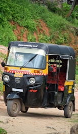 A black and yellow auto rickshaw is parked on a dirt road with lush green foliage in the background. The vehicle has red seating inside and displays signage in a regional script.