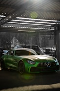 A sleek green sports car is parked in a sunlit garage. The car's glossy paint reflects the light, creating vibrant lens flares. Metal beams and a corrugated roof are visible above, with some trees and a wire fence in the background.